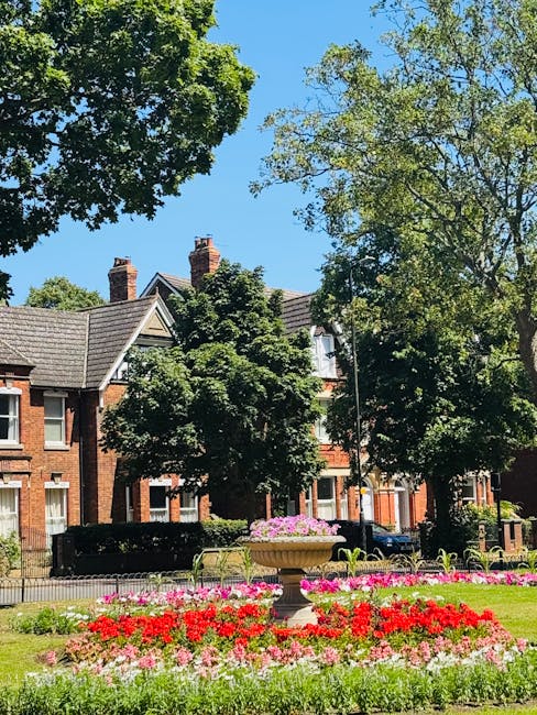 A bright garden scene featuring a well-maintained lawn with neatly trimmed grass and vibrant flower beds arranged in front of a row of traditional red-brick houses. The flower beds display a variety o