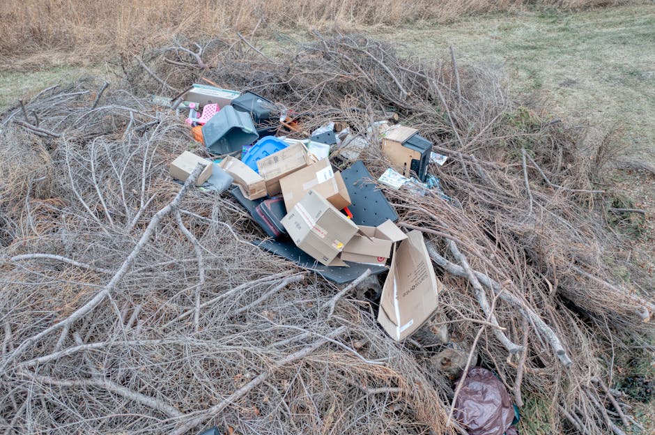 A pile of dry, tangled branches and twigs surrounds a mix of discarded household items, including cardboard boxes, plastic containers, and broken electronic devices, scattered across an outdoor area w