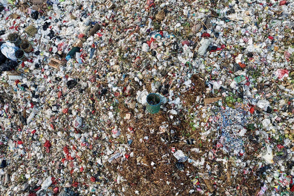 A pile of dry, tangled branches and twigs surrounds a mix of discarded household items, including cardboard boxes, plastic containers, and broken electronic devices, scattered across an outdoor area w
